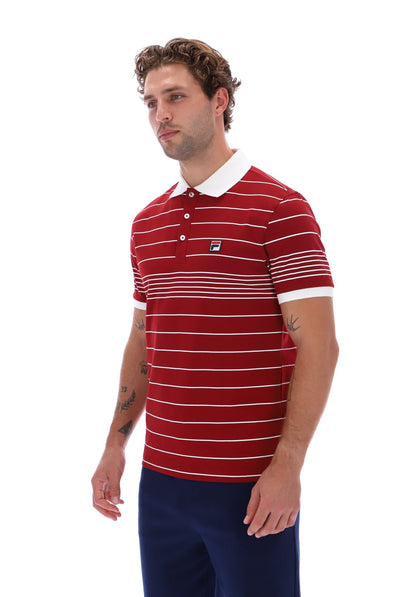 A man wearing a red short-sleeve polo shirt with thin white horizontal stripes, a white collar, and a small Fila logo on the chest, paired with navy blue pants, set against a plain white background.