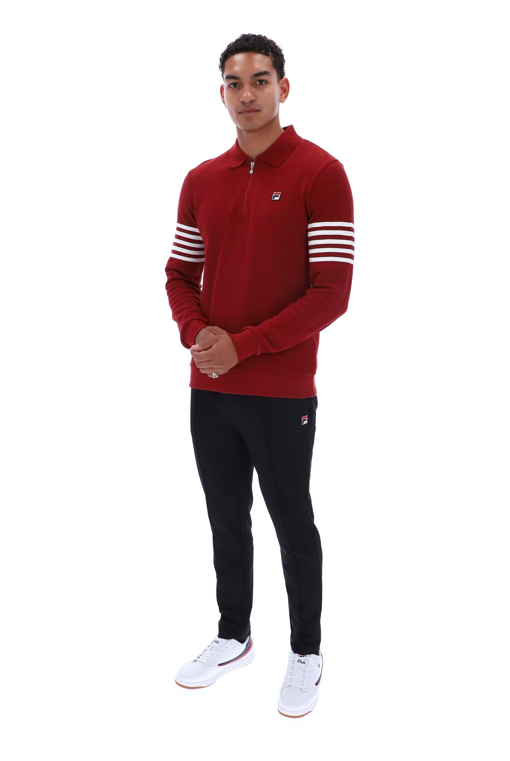 A man modeling a red half-zip pullover sweatshirt with white stripes on the sleeves, paired with black pants and white sneakers, against a plain white background.