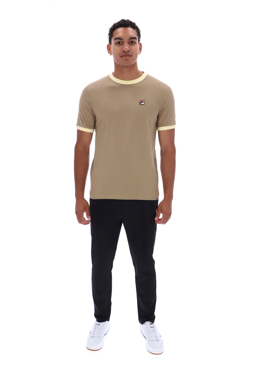 A man modeling a beige ringer t-shirt with yellow trim on the crew neck and sleeve cuffs, paired with black pants and white sneakers, against a plain white background.
