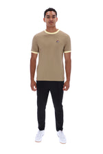 A man modeling a beige ringer t-shirt with yellow trim on the crew neck and sleeve cuffs, paired with black pants and white sneakers, against a plain white background.
