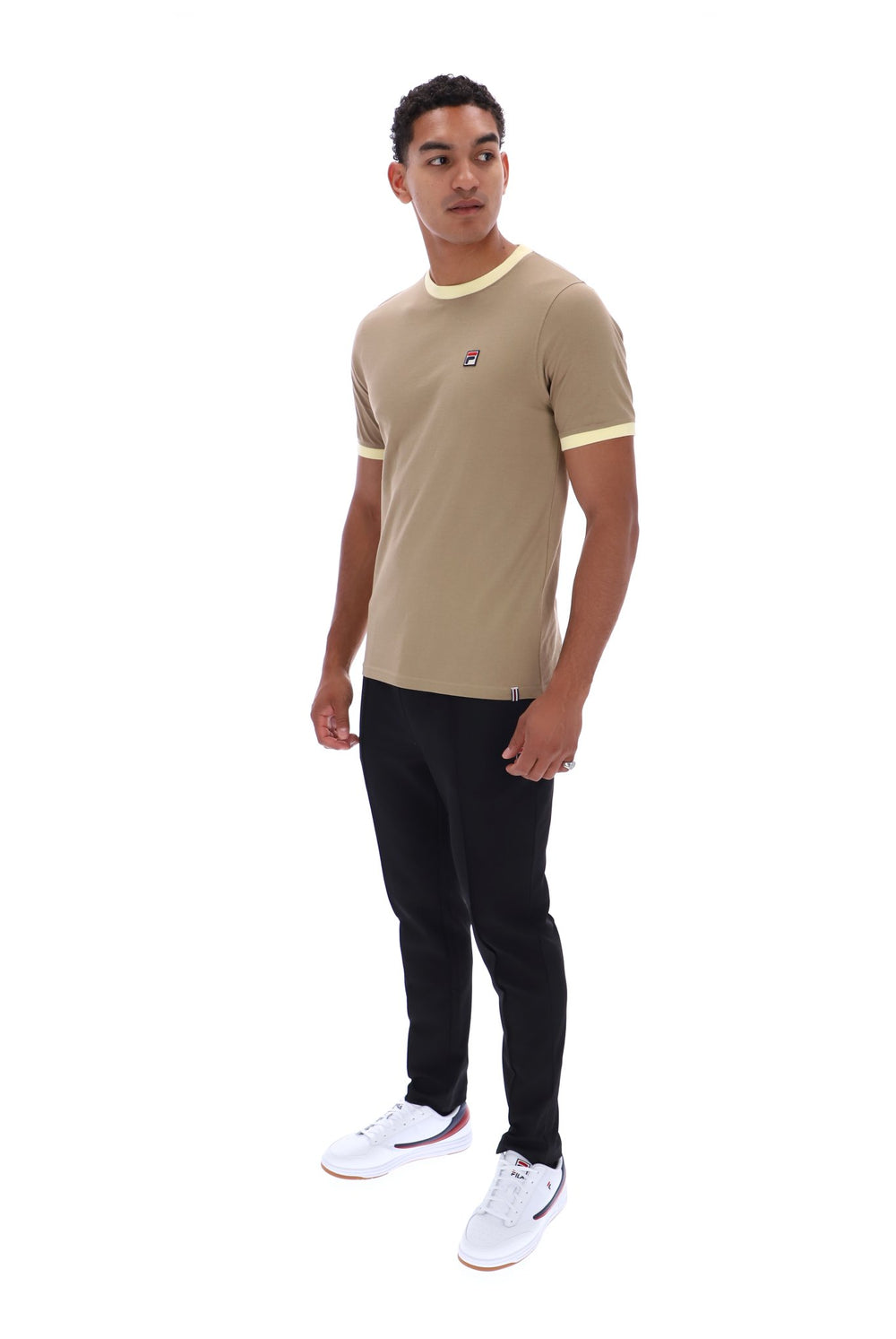 A man modeling a beige ringer t-shirt with yellow trim on the collar and sleeve cuffs, paired with black pants and white sneakers, against a plain white background.