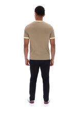 A man viewed from behind wearing a beige short-sleeve t-shirt with light yellow trim on the collar and sleeve cuffs, paired with black pants and white sneakers.