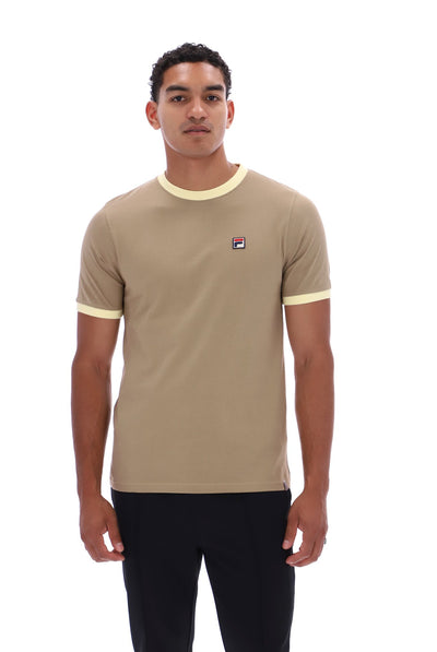 A man wearing a beige ringer t-shirt with cream trim on the collar and sleeve cuffs, featuring a small Fila logo on the left chest, paired with black pants against a white background.