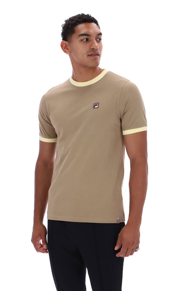 A man wearing a beige ringer t-shirt with cream trim on the collar and sleeve cuffs, featuring a small Fila logo on the left chest, paired with black pants against a white background.