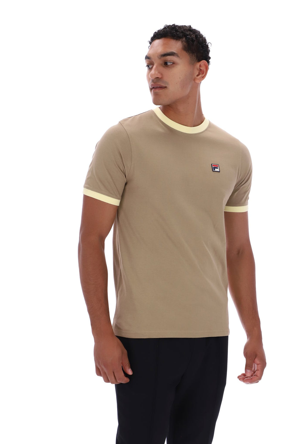 A man wearing a beige ringer t-shirt with cream trim on the collar and sleeve cuffs, featuring a small Fila logo on the left chest, paired with black pants against a white background.