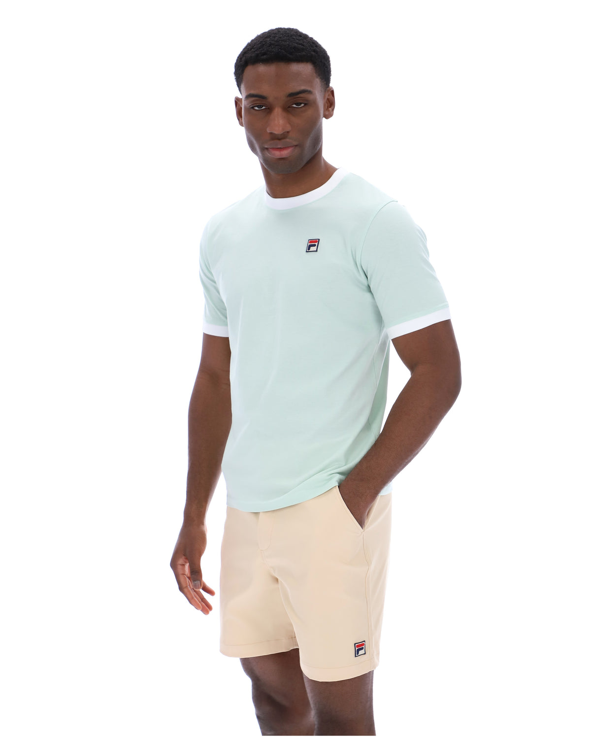 Man wearing a light green t-shirt and beige shorts with a visible brand logo on a white background
