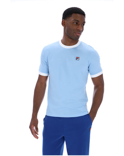 Man wearing a light blue t-shirt with a logo and blue pants on a white background
