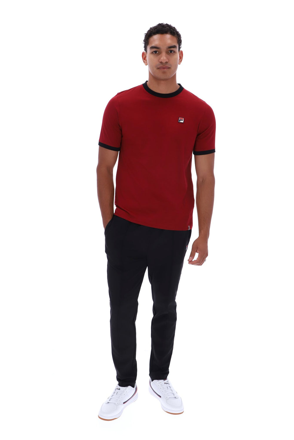 A man modeling a red short-sleeve t-shirt with black trim on the collar and sleeve cuffs, paired with black pants and white sneakers, against a plain white background.