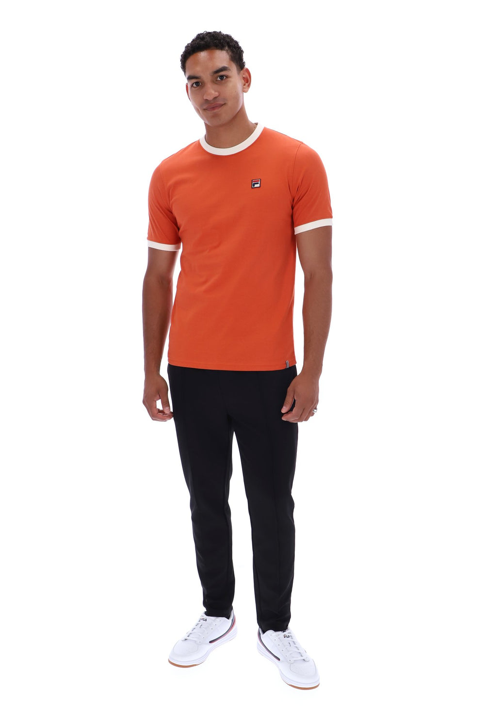 A man modeling a short-sleeve orange t-shirt with white trim on the collar and sleeve cuffs, paired with black pants and white sneakers, against a plain white background.
