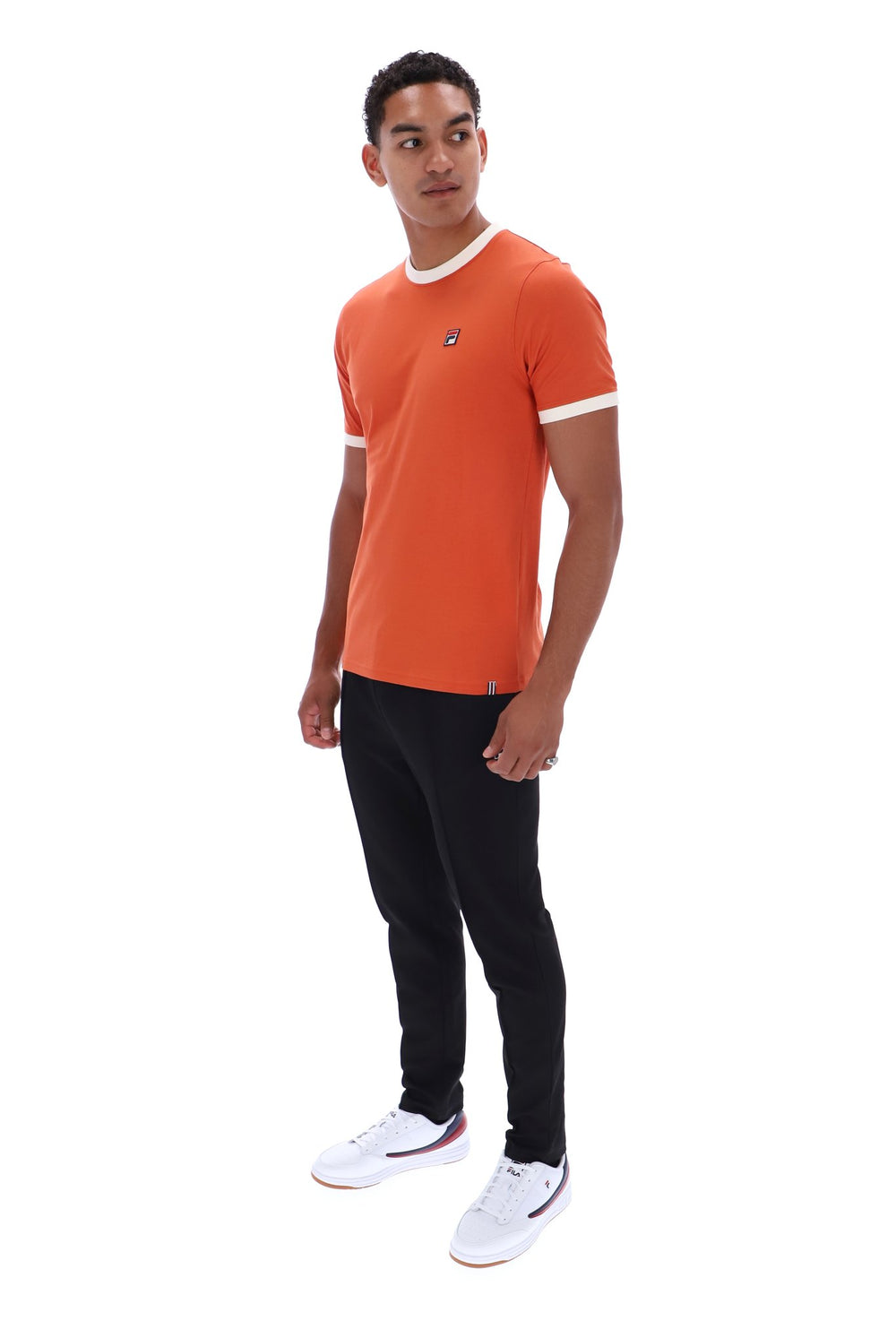 A man modeling a short-sleeve orange t-shirt with white trim on the collar and sleeve cuffs, paired with black pants and white sneakers, against a plain white background.