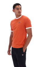 A man wearing a bright orange ringer t-shirt with white trim on the collar and sleeve cuffs, paired with black pants. The shirt features a small embroidered Fila logo on the left chest.