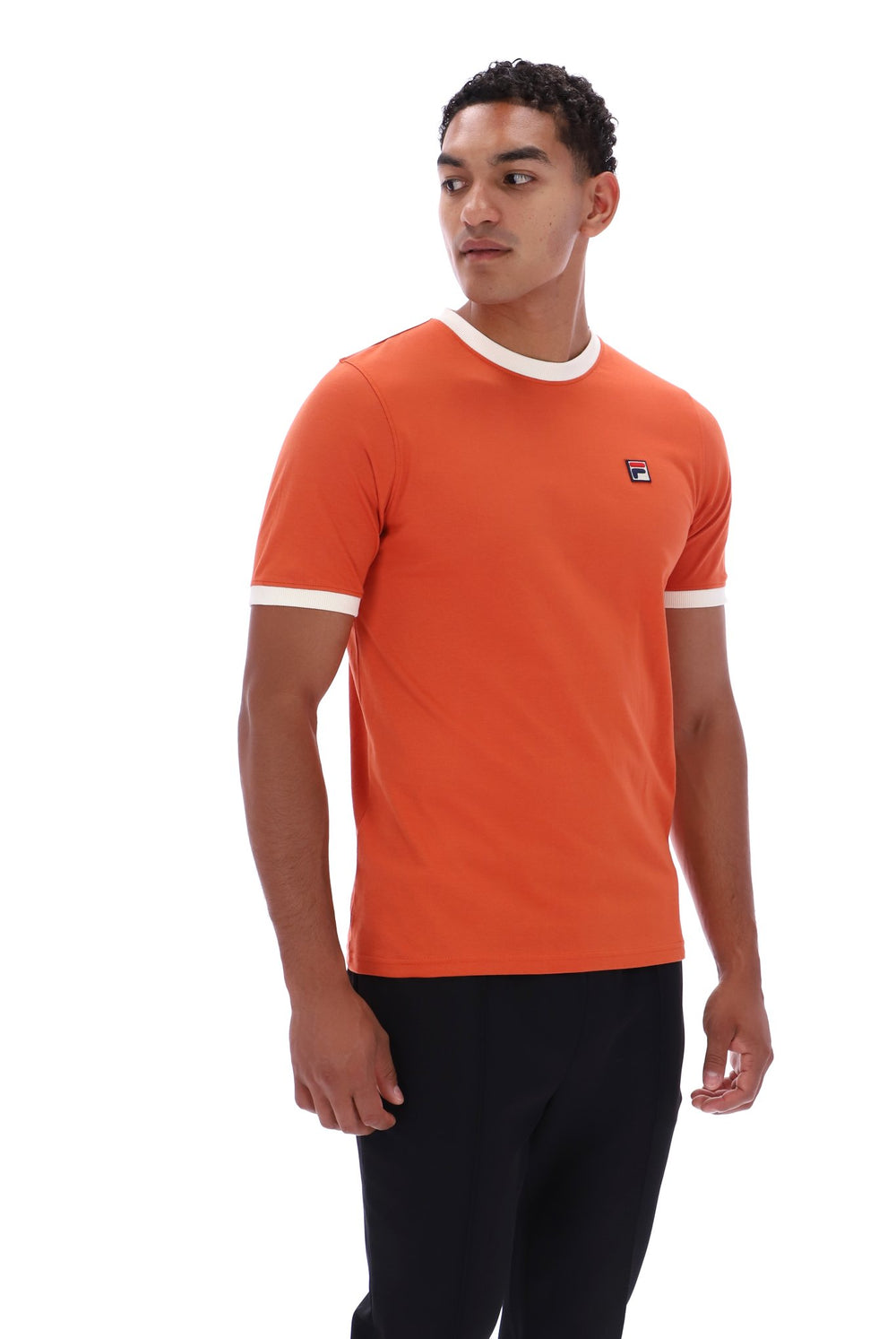 A man wearing a bright orange ringer t-shirt with white trim at the crew neck and sleeve cuffs, paired with black pants, photographed against a solid white background.