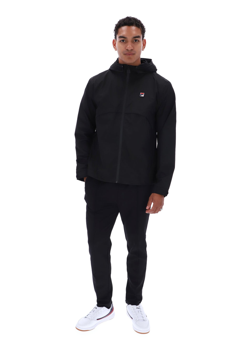 A man modeling a black Fila hooded zip-up jacket, black track pants, and white sneakers against a plain white background.