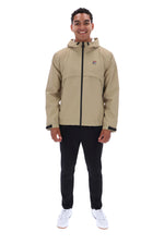 A man wearing a beige hooded jacket with a black zipper, black pants, and white sneakers, standing against a plain white background. The jacket features a small red and white logo on the left chest.