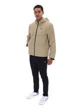 A man modeling a beige Filson hooded jacket with black zippers and a small red logo, paired with black pants and white sneakers, against a plain white background.