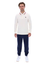 A man modeling a cream long-sleeve polo shirt with a red and blue striped collar and a small Fila logo on the chest, paired with navy joggers and white sneakers, against a plain white background.