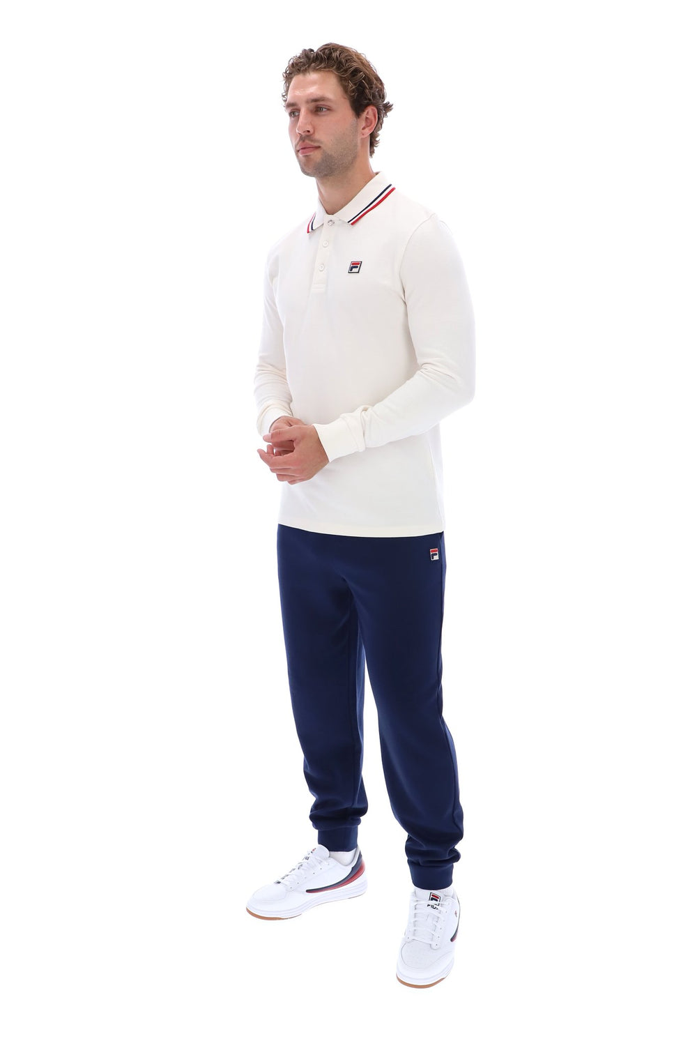 A man modeling a white long-sleeve polo shirt with red and blue striped collar details, navy blue jogger pants, and white sneakers, shown against a plain white background.
