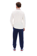 A man viewed from behind wearing a cream long-sleeve polo shirt with red and blue stripes on the collar, navy blue sweatpants, and white sneakers with red and blue accents, against a solid white background.