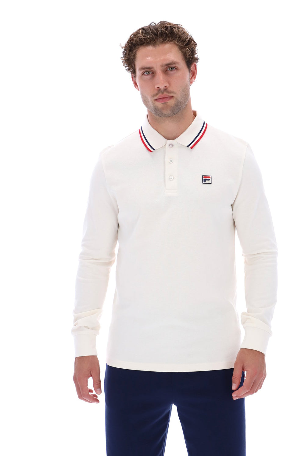 A man wearing a long-sleeve white polo shirt with red and blue striped collar trim and a small Fila logo on the chest, paired with navy pants, against a plain white background.