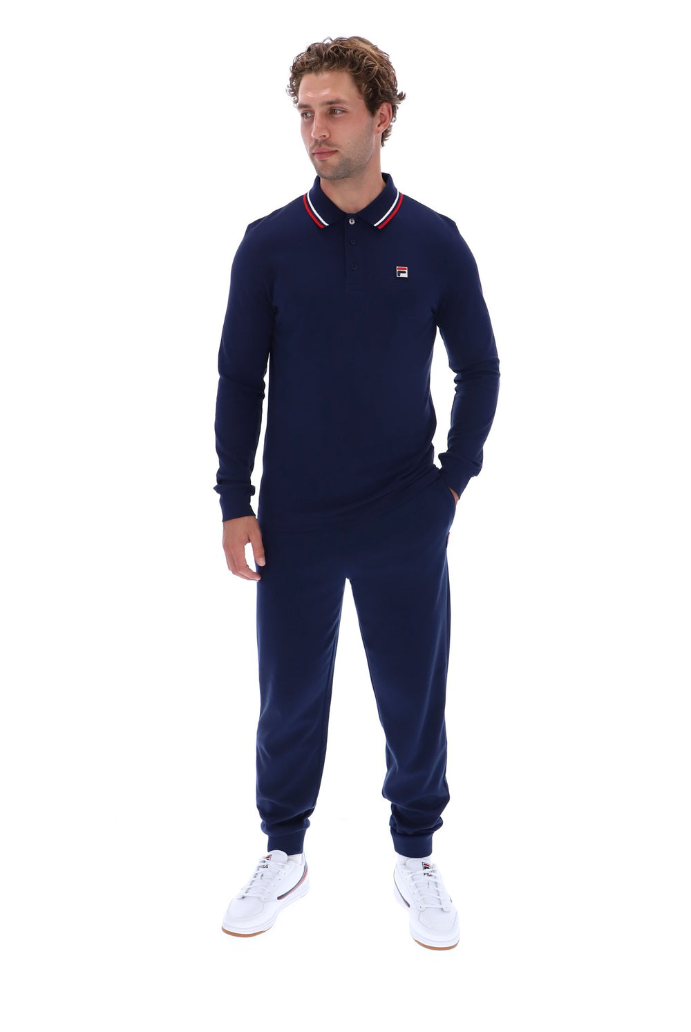 A man modeling a navy blue long-sleeve polo shirt and matching jogger pants set by Fila, paired with white sneakers, against a solid white background.