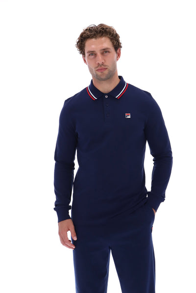 A man wearing a navy blue long-sleeve polo shirt with a red, white, and blue striped collar and a small Fila logo on the chest, paired with matching navy pants, against a solid white background.
