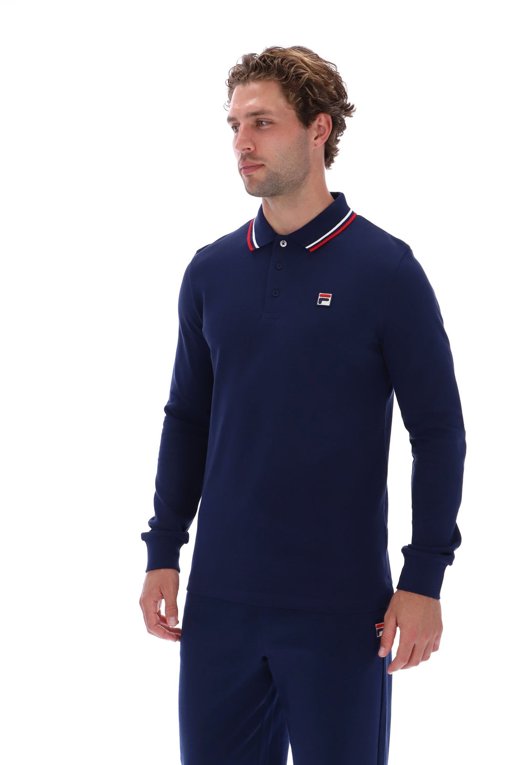 A man wearing a navy blue long-sleeve polo shirt with a red and white striped collar and a small Fila logo on the chest, paired with matching navy pants, against a plain white background.