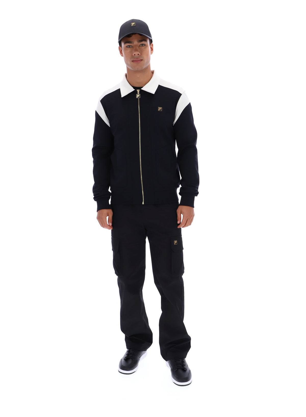 Man wearing a black and white jacket with a logo, black pants, and a cap on a white background