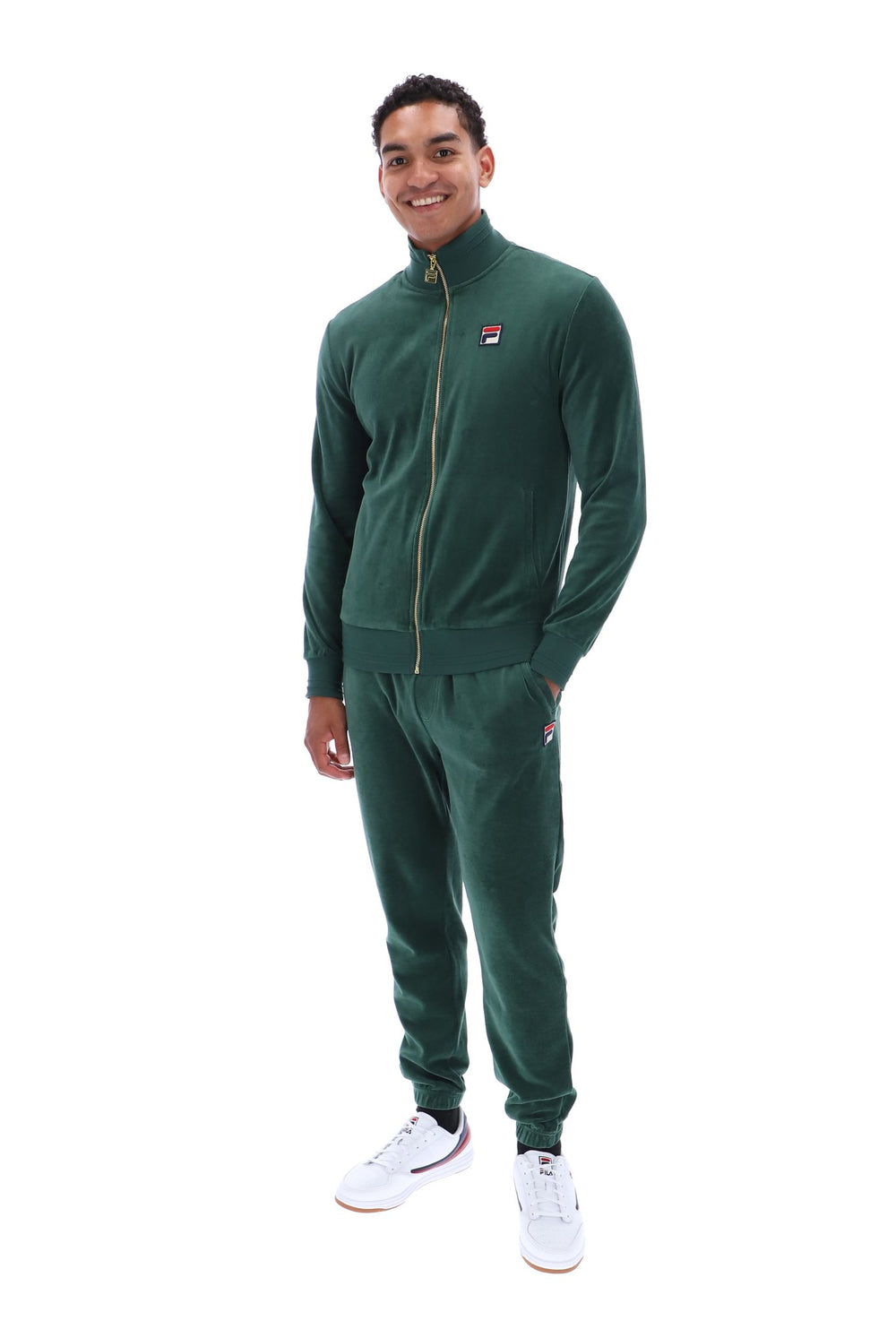 A man modeling a dark green Fila tracksuit, featuring a zip-up jacket with a small red, white, and blue logo on the left chest, and matching jogger pants, against a plain white background.