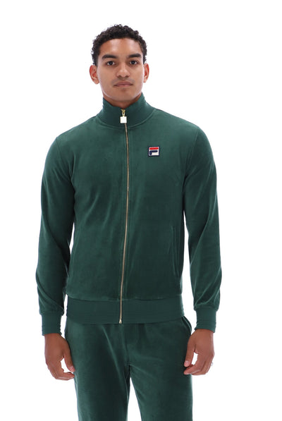A man wearing a dark green Fila velour tracksuit with a matching zip-up jacket and pants, featuring a small red, white, and blue logo on the chest, set against a plain white background.