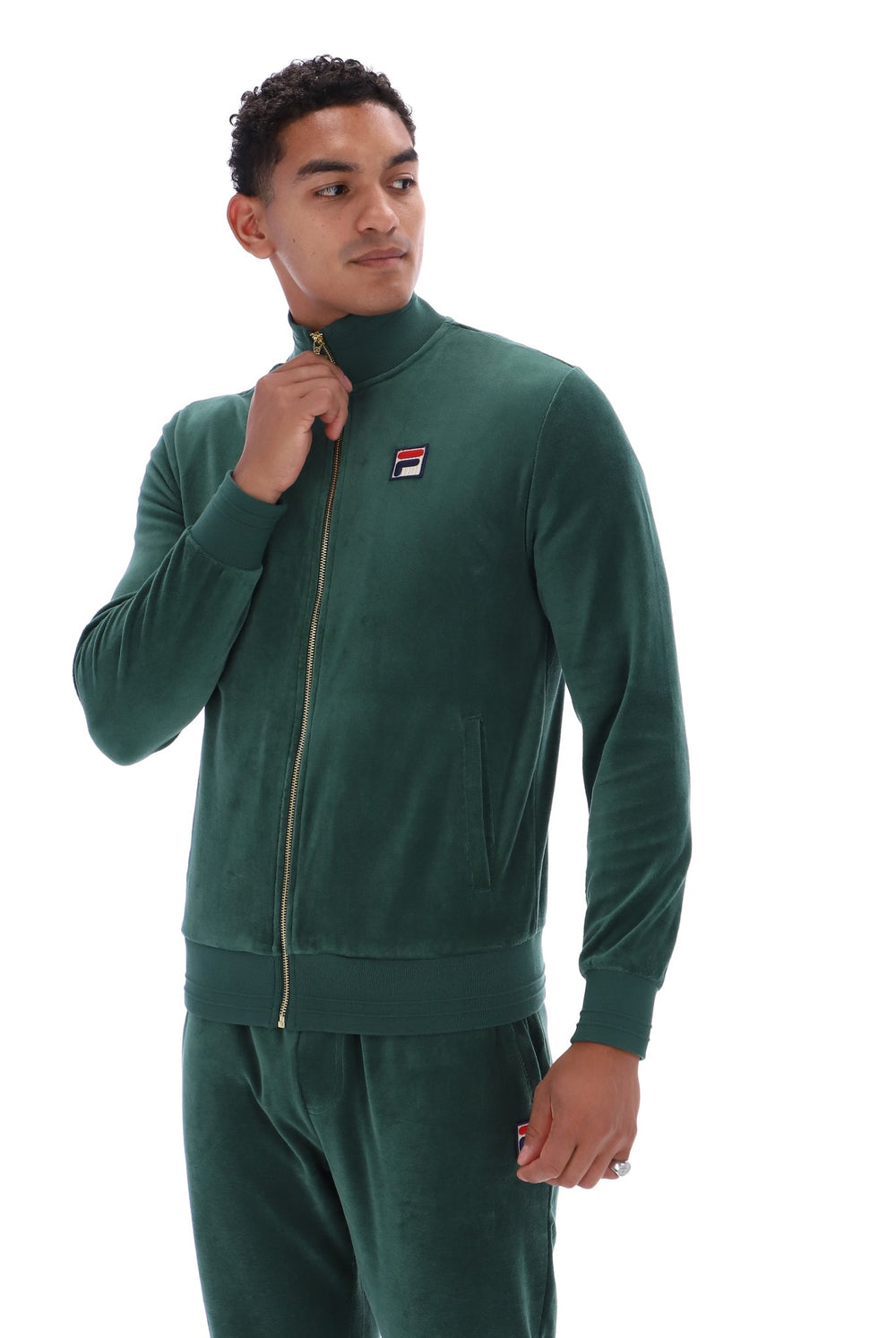 A man wearing a dark green velour tracksuit with a matching zip-up jacket and pants, featuring a small Fila logo patch on the left chest, set against a plain white background.