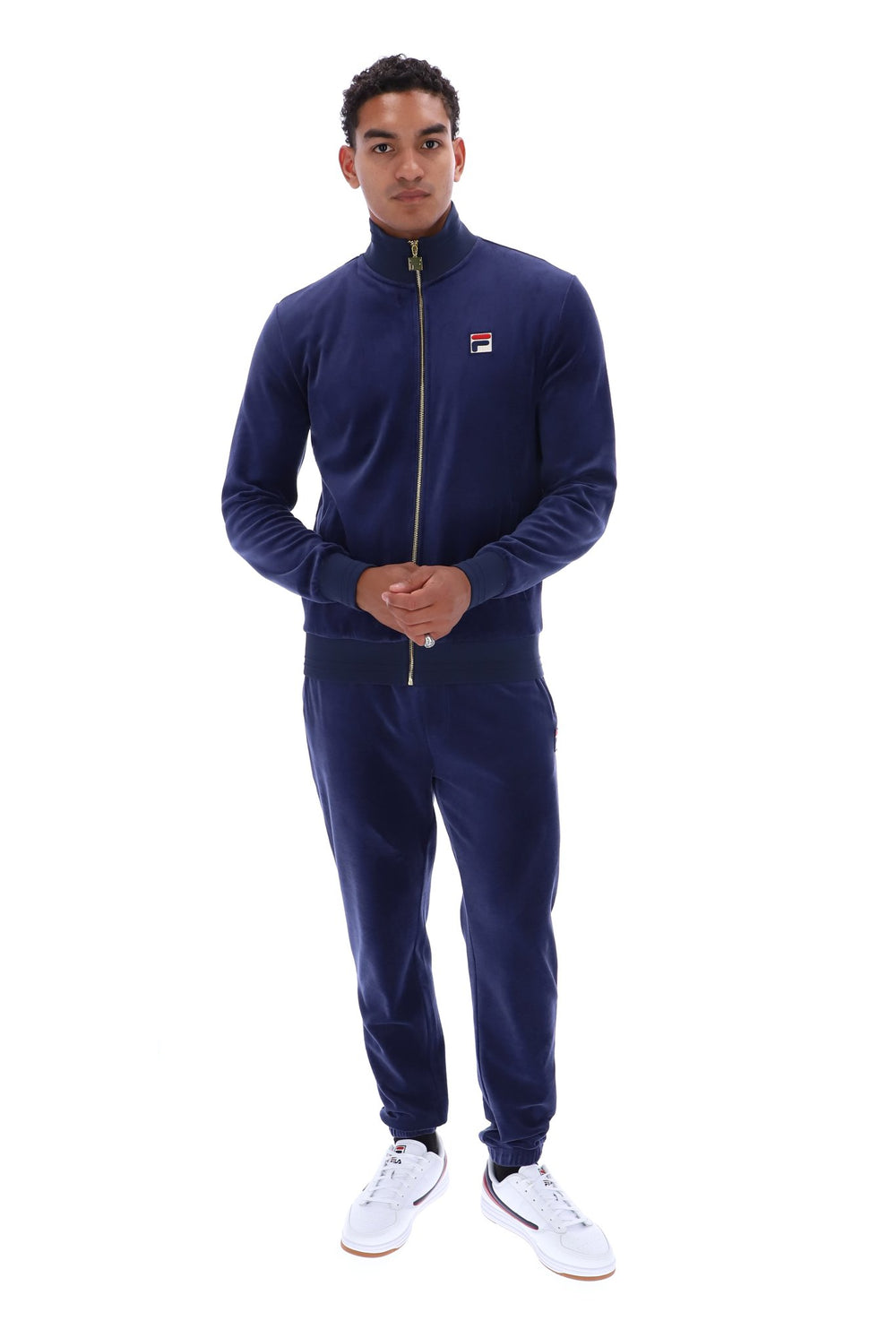 A man modeling a navy blue Fila zip-up track jacket and matching sweatpants set, paired with white sneakers, against a solid white background.