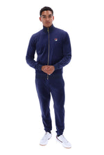 A man modeling a navy blue Fila zip-up track jacket and matching sweatpants set, paired with white sneakers, against a solid white background.