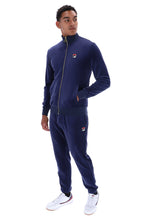 A man modeling a navy blue Fila tracksuit, featuring a zip-up jacket and matching jogger pants, with small red, white, and blue logo patches on the chest and thigh. He is wearing white sneakers against a plain white background.