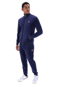 A man modeling a navy blue Fila tracksuit, featuring a zip-up jacket and matching jogger pants, with small red, white, and blue logo patches on the chest and thigh. He is wearing white sneakers against a plain white background.