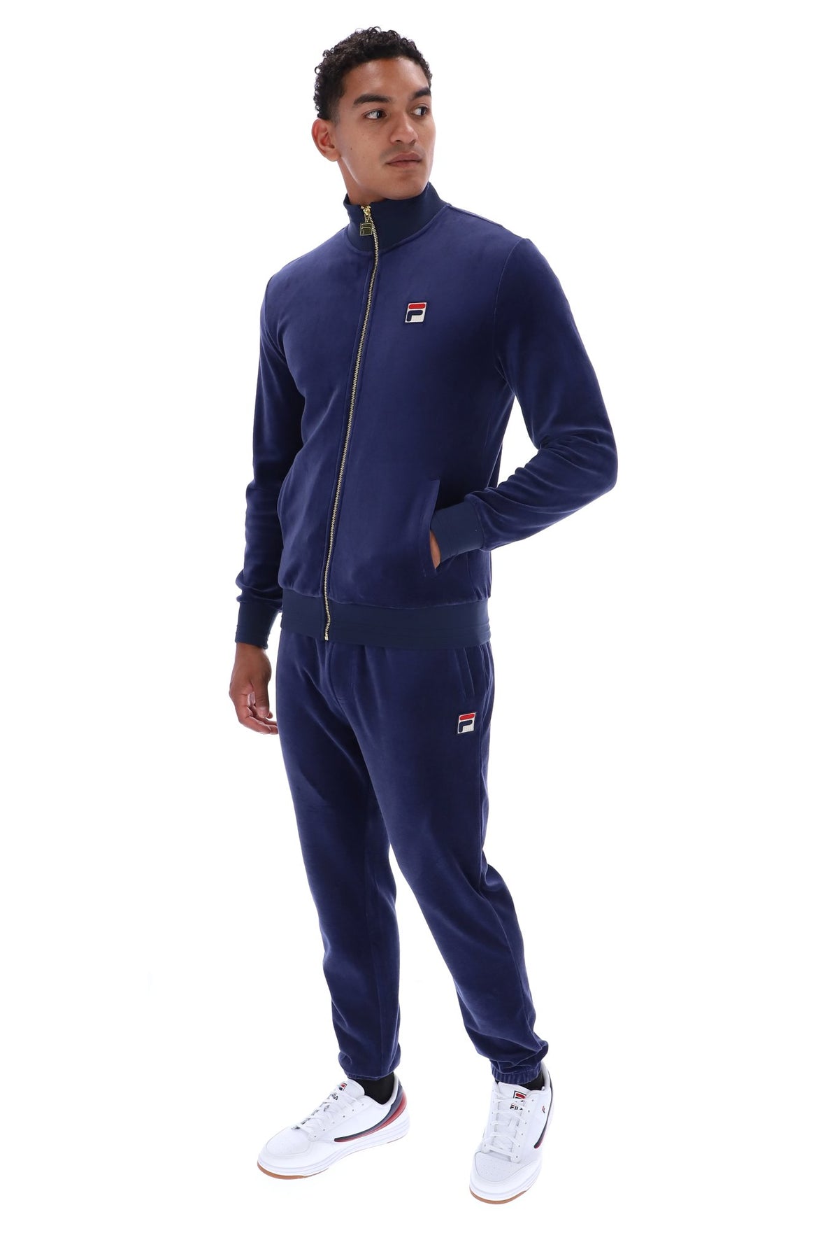 A man modeling a navy blue Fila tracksuit, featuring a zip-up jacket and matching jogger pants, with small red, white, and blue logo patches on the chest and thigh. He is wearing white sneakers against a plain white background.