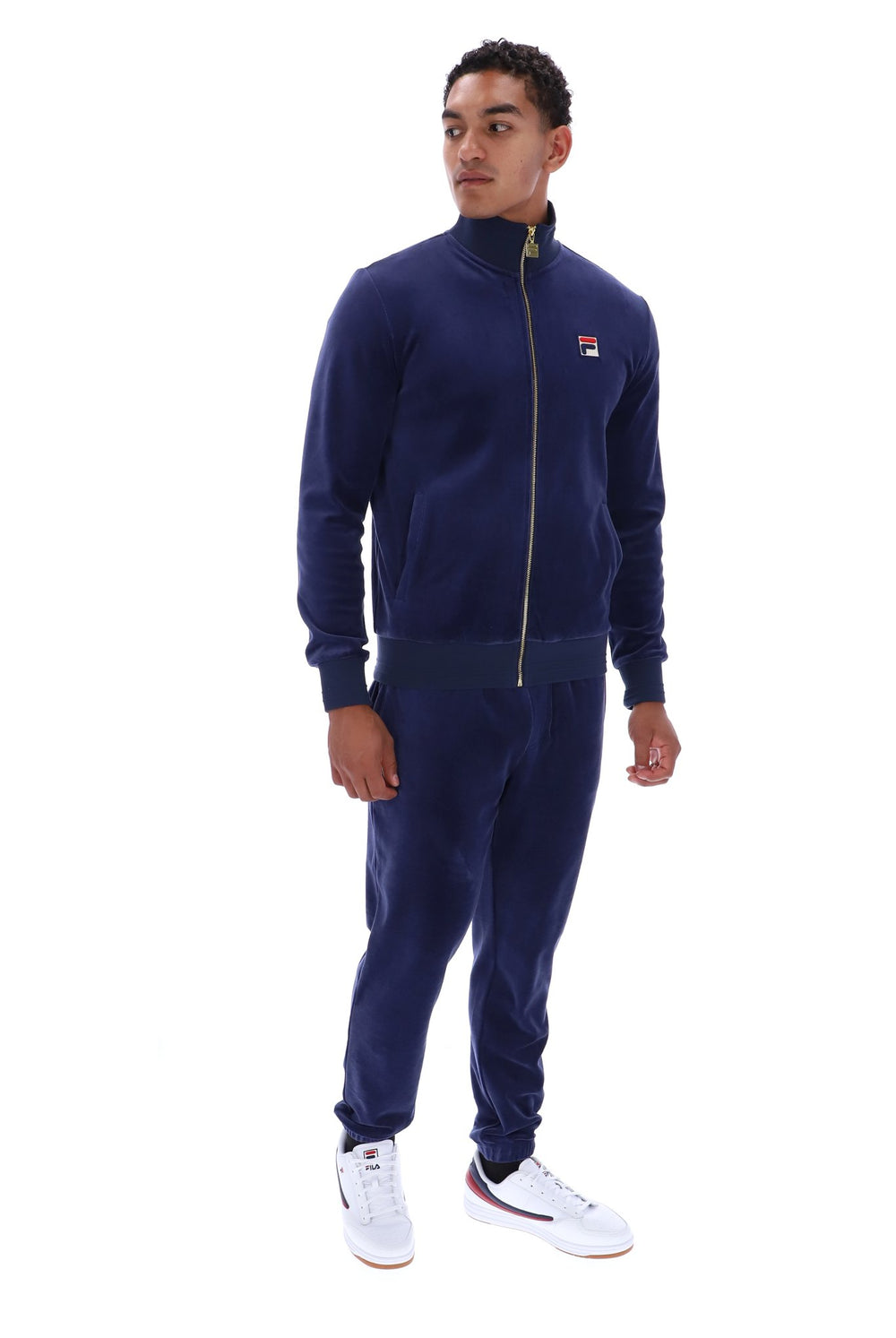 A man modeling a navy blue tracksuit with a zip-up jacket and matching joggers, featuring a small red, white, and blue Fila logo on the left chest, paired with white sneakers against a plain white background.