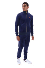 A man modeling a navy blue tracksuit with a zip-up jacket and matching joggers, featuring a small red, white, and blue Fila logo on the left chest, paired with white sneakers against a plain white background.