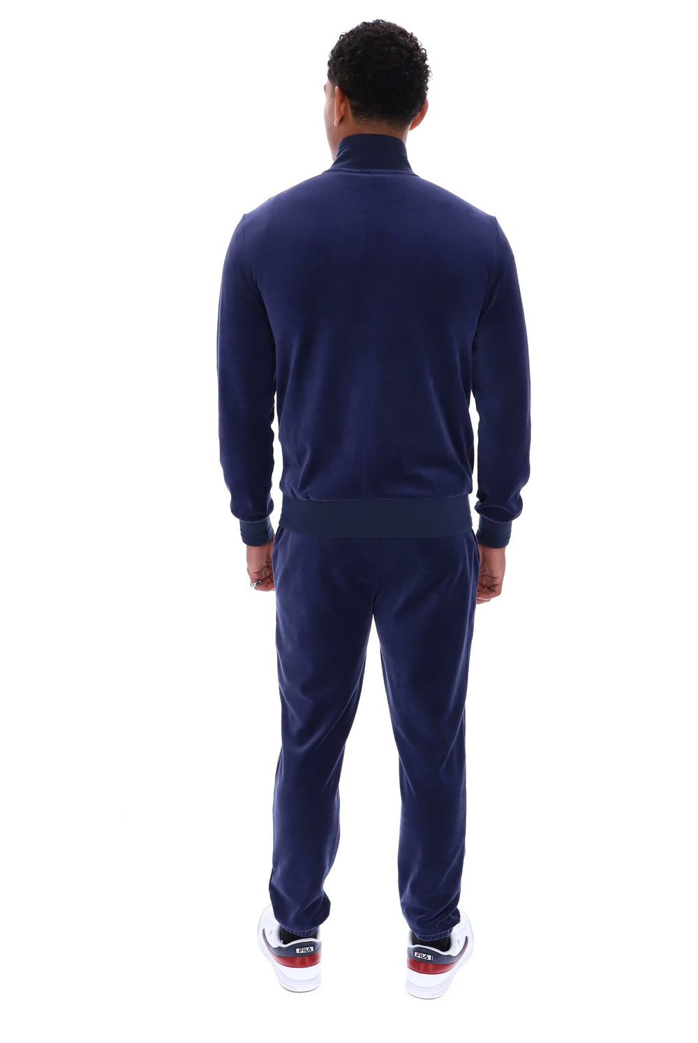 A man viewed from the back wearing a navy blue velour tracksuit with a mock neck jacket and matching pants, paired with white sneakers.