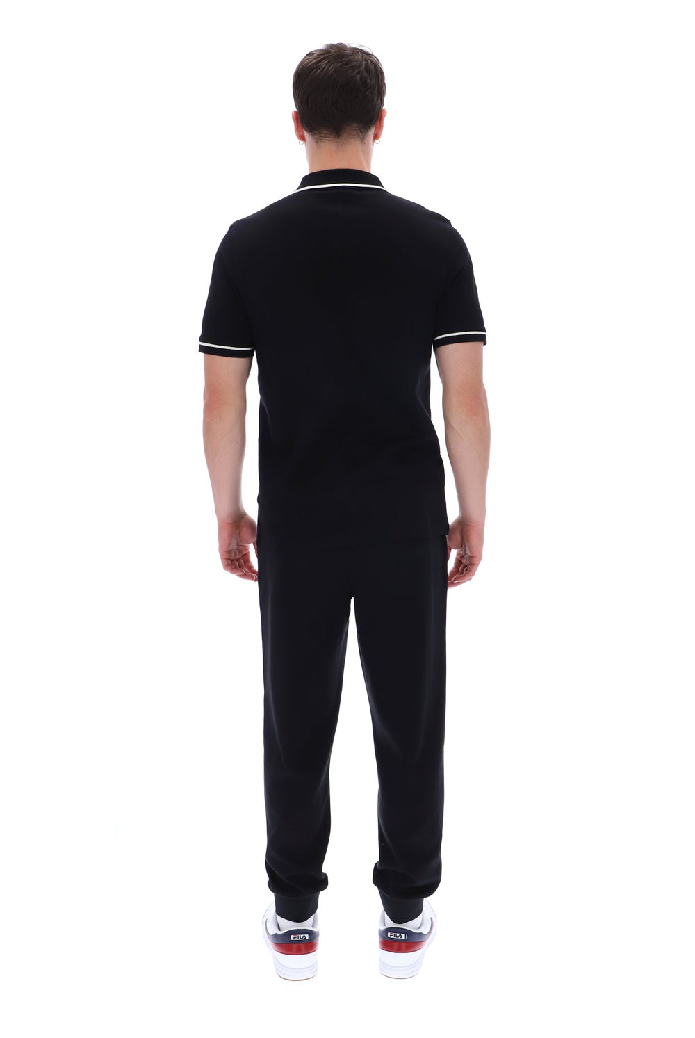 A man viewed from behind wearing a black short-sleeve polo shirt with white trim on the collar and sleeve cuffs, paired with black sweatpants and white sneakers with red accents, against a plain white background.