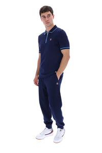 A man modeling a navy blue polo shirt and matching sweatpants set with white sneakers, photographed against a solid white background.