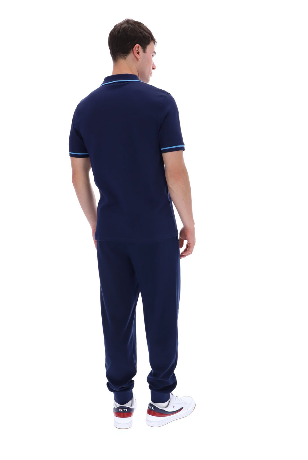 A man shown from the back wearing a navy blue short-sleeve polo shirt with light blue and white trim on the collar and sleeve cuffs, matching navy jogger pants, and white sneakers with red accents, set against a plain white background.