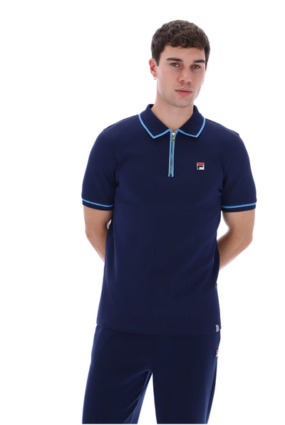 A man wearing a navy blue short-sleeve polo shirt with light blue trim and a small Fila logo on the chest, paired with matching navy pants, against a plain white background.