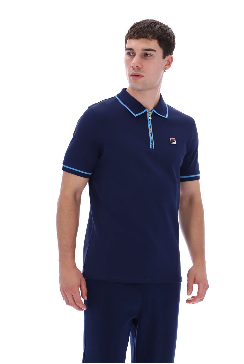 A man wearing a navy blue short-sleeve polo shirt with light blue trim on the collar, sleeve cuffs, and a small red and white Fila logo on the left chest, paired with matching navy pants, against a plain white background.