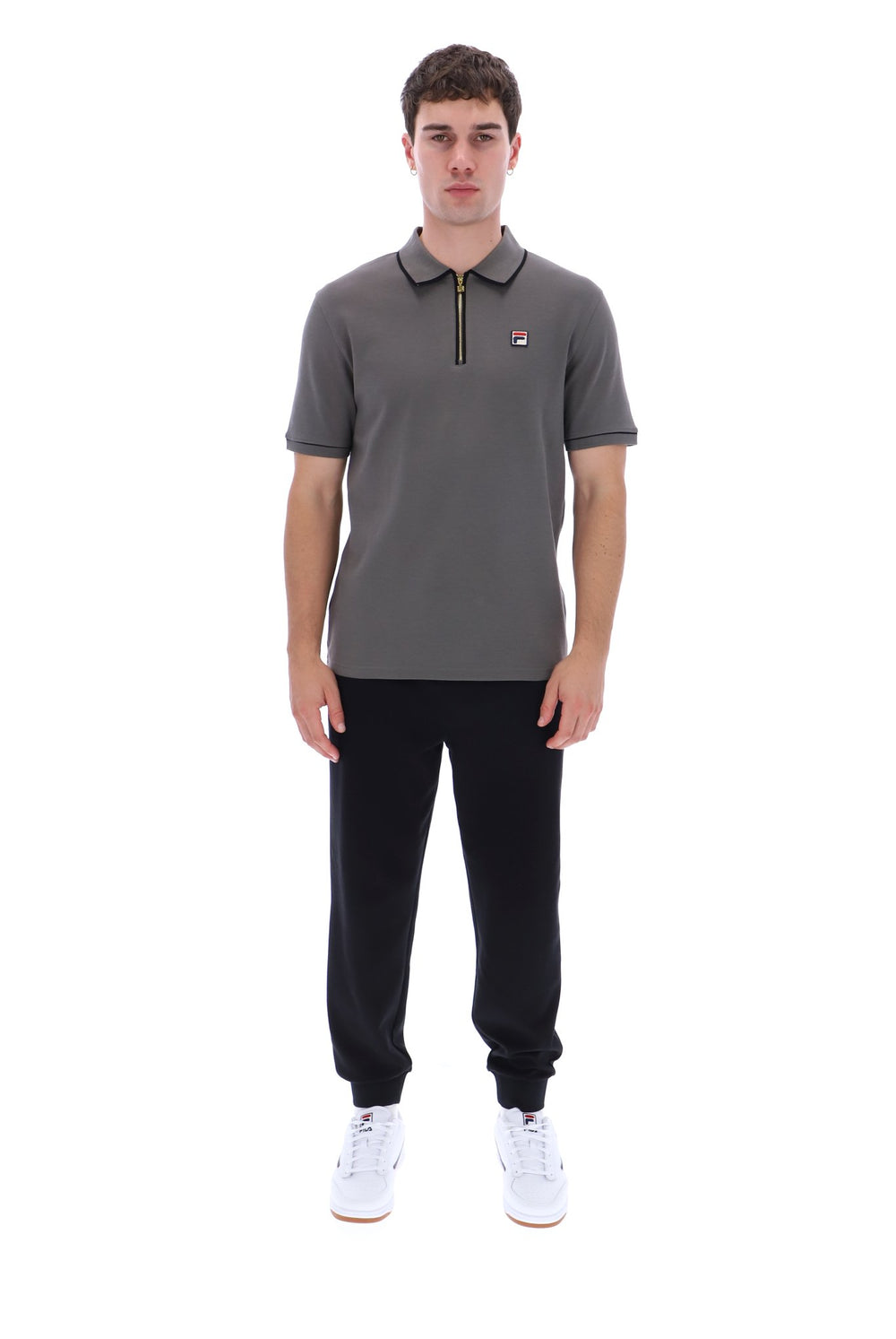A man modeling a dark gray short-sleeve polo shirt with a half-zip collar, black jogger pants, and white sneakers against a plain white background.