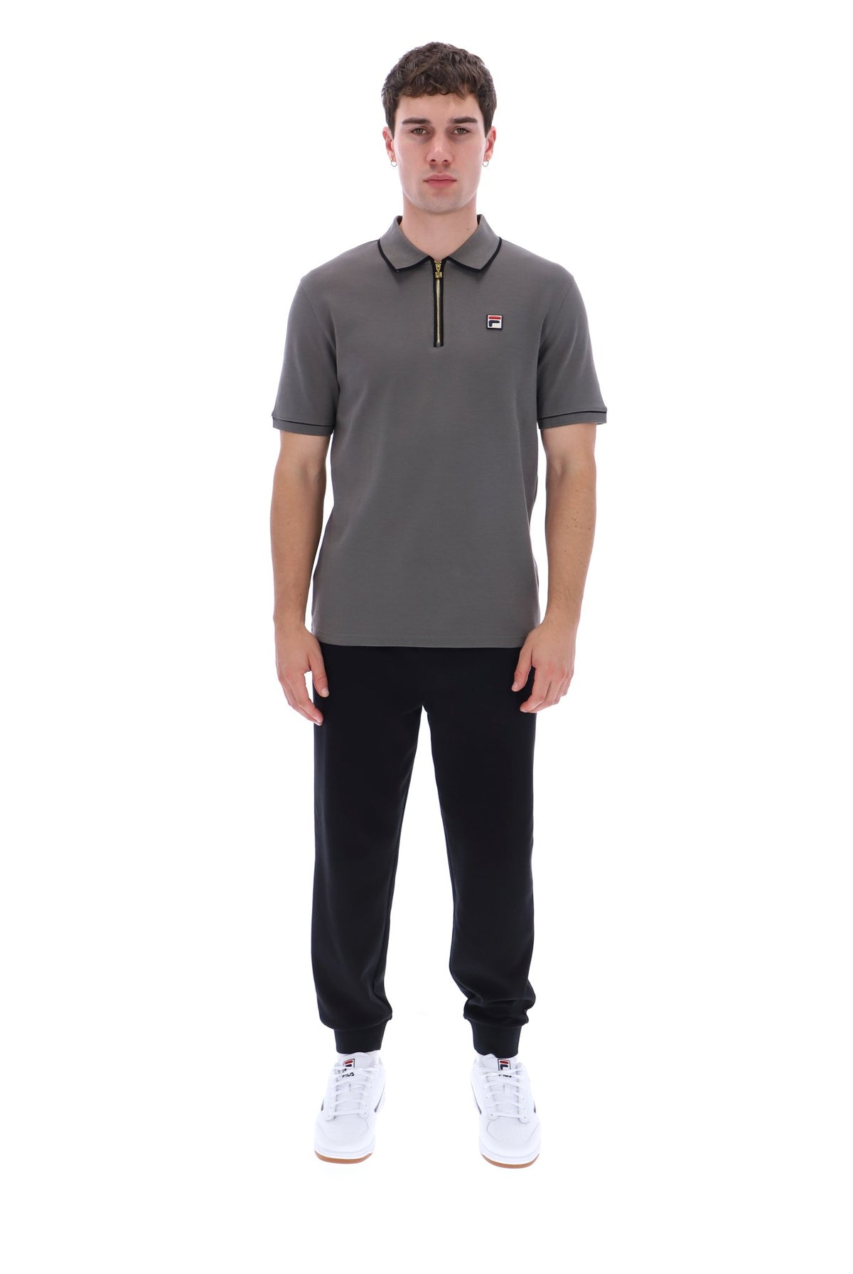A man modeling a dark gray short-sleeve polo shirt with a half-zip collar, black jogger pants, and white sneakers against a plain white background.