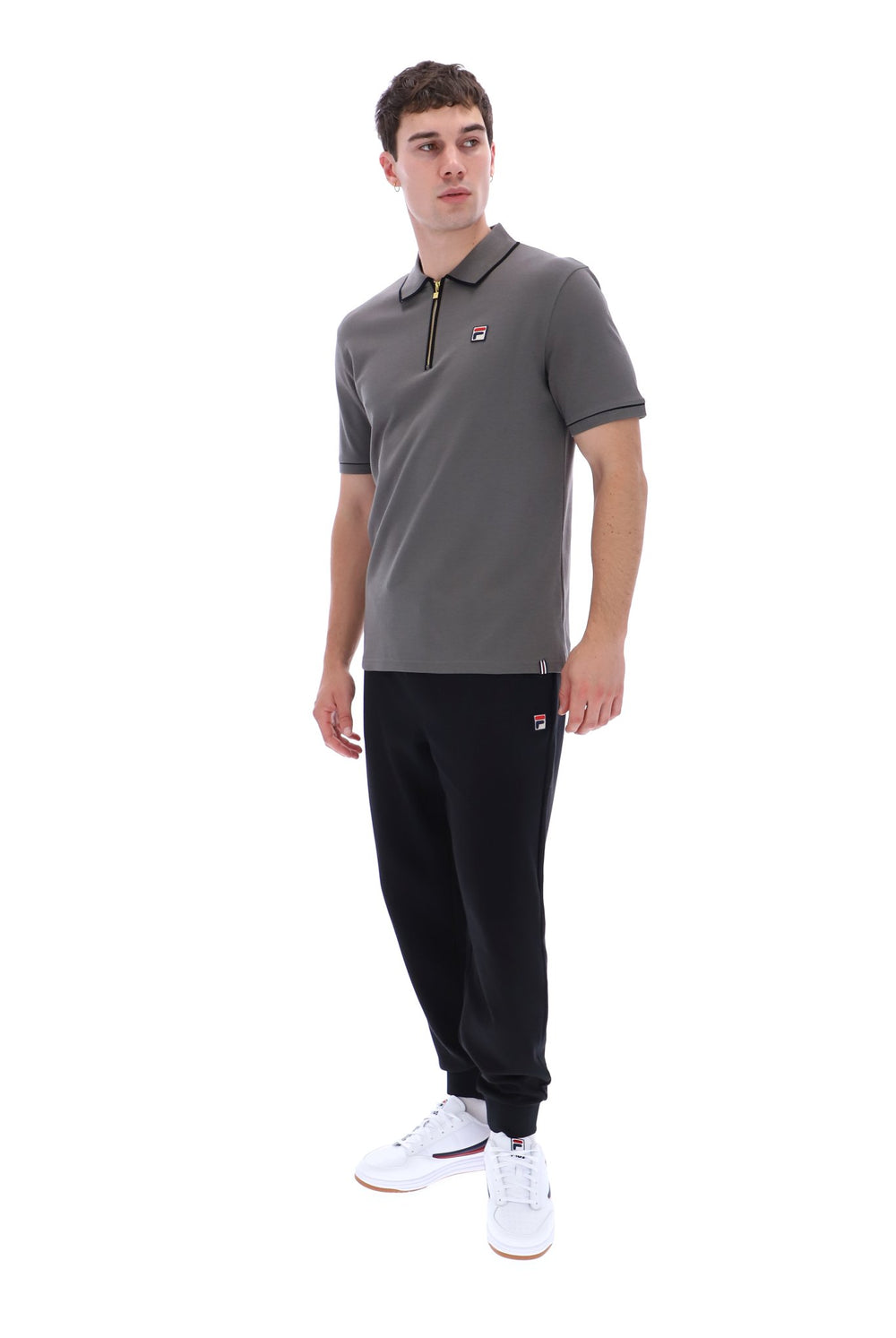 A man modeling a gray short-sleeve polo shirt with a black zipper and a small red and white logo on the chest, paired with black joggers and white sneakers, against a plain white background.