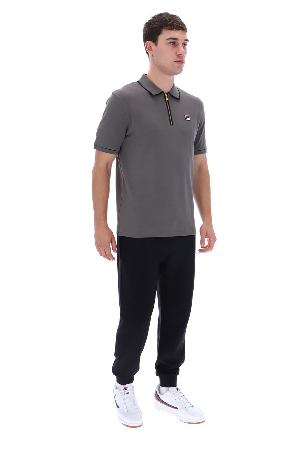 A man modeling a gray short-sleeve polo shirt with a black zipper collar, paired with black joggers and white sneakers, shot against a solid white background.