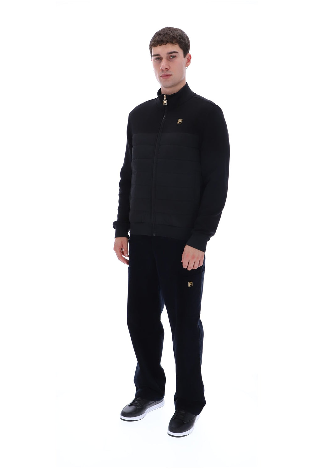A man modeling a black zip-up jacket and matching black trousers with small gold embroidered logo details, photographed against a solid white background.