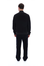 A man viewed from behind wearing a black long-sleeve knit top and matching black trousers, standing against a plain white studio background.