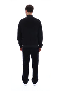 A man viewed from behind wearing a black long-sleeve knit top and matching black trousers, standing against a plain white studio background.